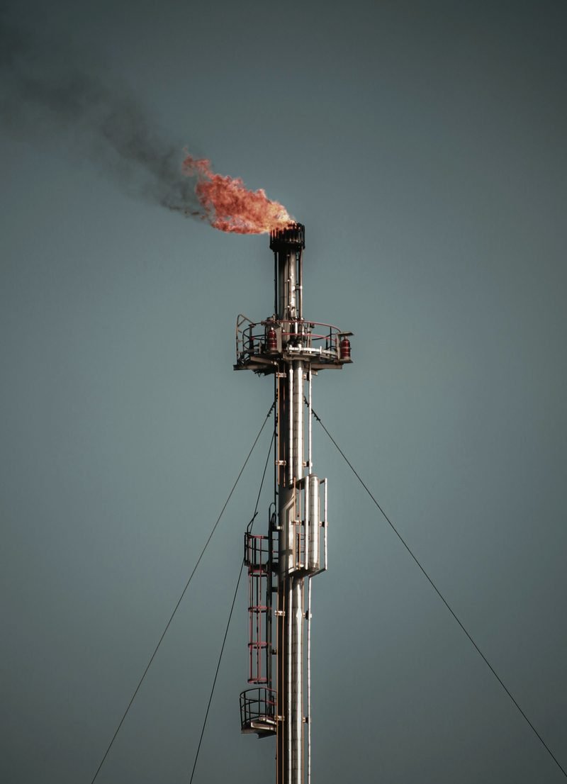 A tall steel flare stack burning a small orange flame against a grey-blue sky, held by guy wires.