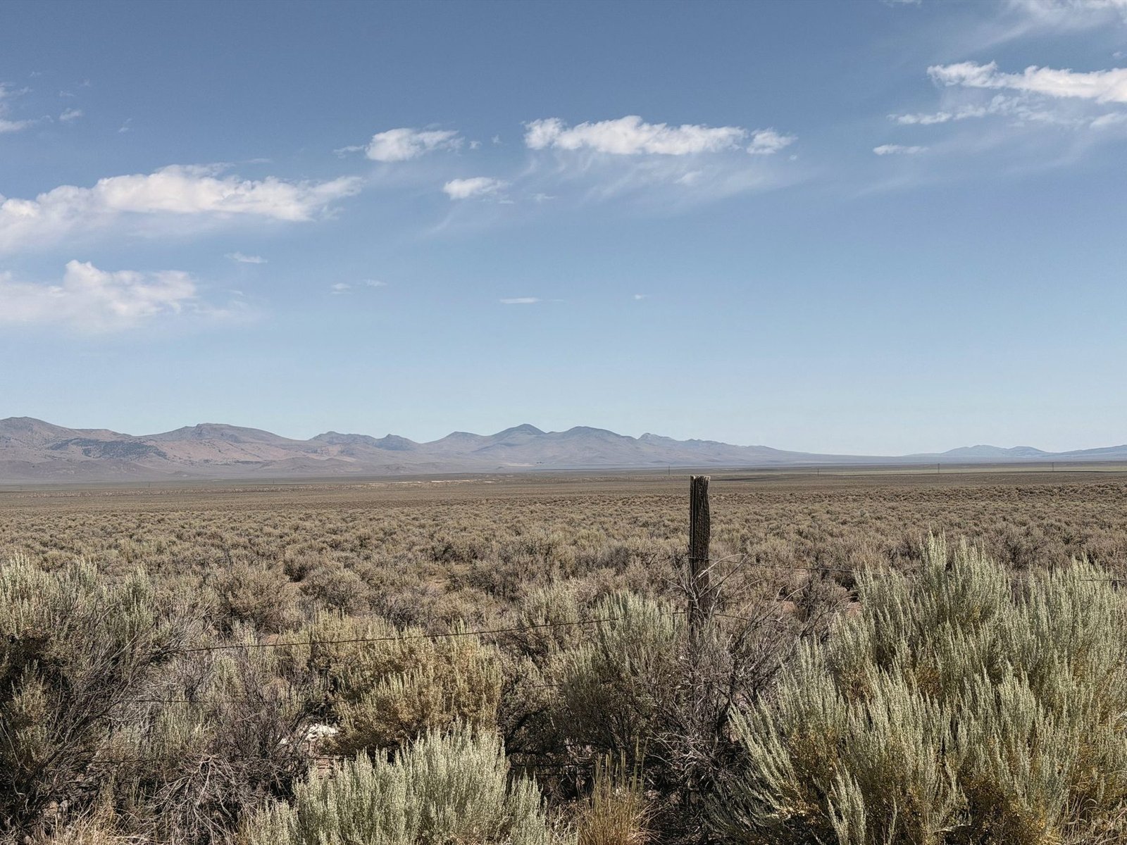 Sagebrush flats and a single fence post under a wide blue sky, with bare mountains on the horizon.